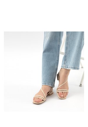 Stopy w beżowych sandałach na niskim obcasie z krzyżującymi się paskami, w połączeniu z jasnoniebieskimi, prostymi jeansami, siedzące na białym krześle.