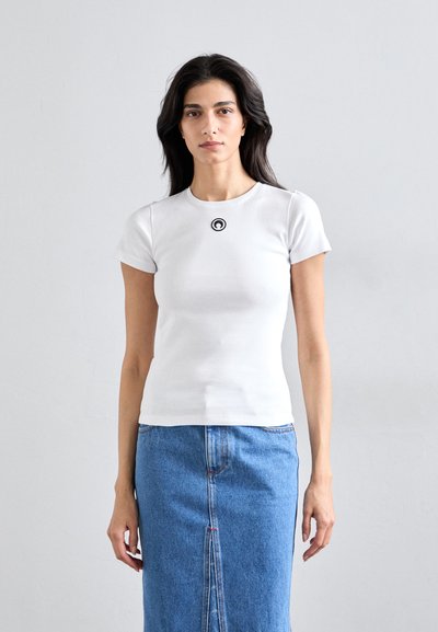 T-shirt ajusté à manches courtes blanc avec un logo graphique noir sur la poitrine, associé à une jupe en denim bleu, avec une fente sur le devant et des coutures visibles.