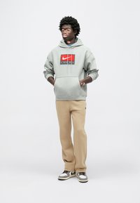 Γκρι φούτερ με κουκούλα με μαύρο και κόκκινο λογότυπο Nike, μπεζ φόρμα και γκρι αθλητικά παπούτσια με μαύρες και λευκές λεπτομέρειες. Καθημερινή, άνετη γραμμή.