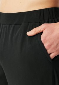 Zwarte sportbroek van gladde stof met een elastische tailleband en zijzakken, getoond op heuphoogte met een hand in een zak.