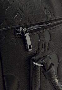 Gros plan sur un sac en cuir noir avec des visages de Mickey Mouse en relief, une tirette de fermeture éclair en argent et une sangle en cuir avec une boucle en métal.