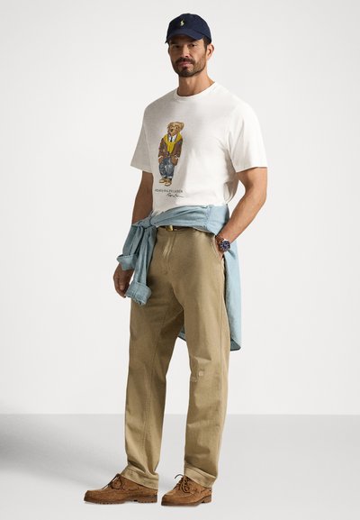 Ανδρικό ανοιχτόχρωμο κρεμ T-shirt με γραφικό αρκούδας, χακί παντελόνι και ανοιχτό μπλε πουκάμισο δεμένο στη μέση. Καφέ σουέντ παπούτσια ολοκληρώνουν το σύνολο.