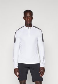 Hvit langermet treningstrøye med halv-zip krage, svarte skulderdetaljer, og "Peak Performance"-logo på høyre erm.