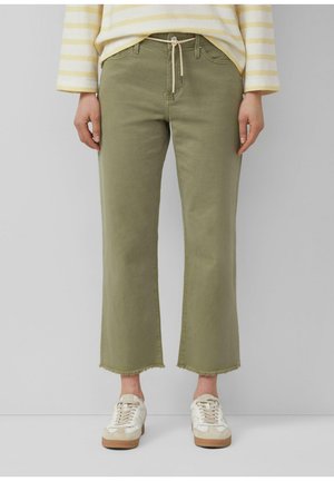 Osoba ubrana w oliwkowo-zielone, skrócone spodnie o szerokich nogawkach z surowymi krawędziami, białe trampki oraz żółto-pasiasty, długi rękaw, stojąca na tle szarego tła.