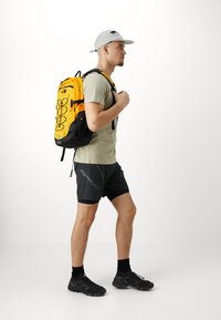 Sac à dos jaune avec des accents noirs, cordon élastique à l'avant et bretelles réglables. Porté avec un tee-shirt gris et un short noir.