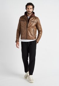 Ruskea nahkatakki, jossa on vetoketjulliset taskut, tekstuurilla koristellut olkapäät ja ruskea huppari alta. Yhdistetty mustiin housuihin ja valkoisiin tennareihin.