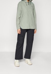 Giacca a zip verde chiaro con cappuccio, progettata con una vestibilità rilassata. Abbinata a pantaloni cargo scuri e scarpe da ginnastica chiare.