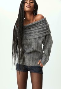 Grauer schulterfreier Pullover mit geripptem Muster und breitem umgeschlagenem Kragen, kombiniert mit dunklen Jeans-Shorts und durchsichtigen schwarzen Strumpfhosen.