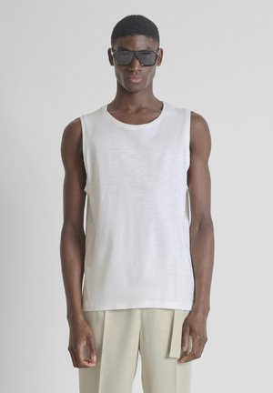 Jeune homme portant de grandes lunettes de soleil noires, un débardeur blanc et un pantalon beige, debout devant un fond gris clair uni.