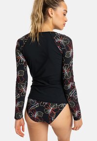 Černý surfovací top s dlouhým rukávem a vzorovanými květinovými rukávy; odpovídající bikiny mají stejný květinový design a kontrastní černé prvky.