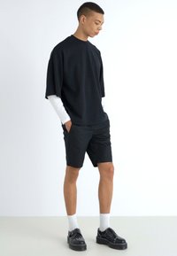 Svart överdimensionerad kortärmad skjorta lagd över en vit långärmad skjorta, tillsammans med svarta shorts och svarta skor med vita strumpor.