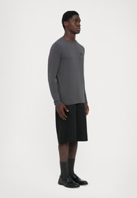 Man standing wearing gray long-sleeve shirt, black knee-length shorts, dark socks, and black loafers against a plain white background.