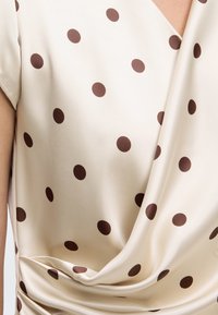 Blouse en satin crème à pois marron, avec un col drapé et des manches courtes.