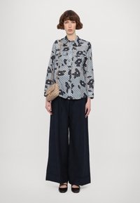 Dlouhý rukáv košile v světle modré barvě s tmavými květinovými vzory a strukturovaným pozadím. V kombinaci s širokými tmavými kalhotami a malou béžovou kabelkou.