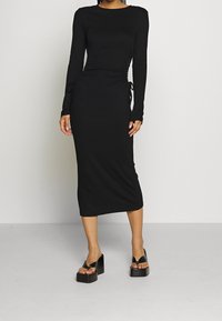 Musta pitkähihainen mekko, jossa on tiukka siluetti, ryhmitellyt sivut ja solmitysdetail. Sujuva kangas ja nilkkapituiset helma. Parit paksupohjaisia mustia sandaaleja.
