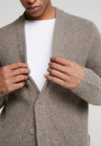 Brunstickad cardigan med ribbad textur, öppen front och tre knappar. Bärs över en vit rundhalsad tröja, vilket framhäver dess varma material.