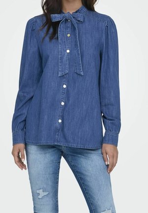 Femme portant un chemisier en jean bleu à manches longues, boutonné avec un nœud noué au col et un jean déchiré bleu clair.