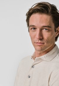 Ljust beige polotröja med en mjuk textur, med en krage och treknapps knäppning, bärs av en person med kort, rufsigt hår.