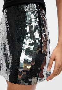 Saia mini de lantejoulas pretas e prateadas em uma pessoa, com a mão e parte da coxa visíveis, mostrando o detalhe do fecho na lateral.