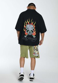 Camicia nera oversize con una grafica di teschio e fiamme sulla schiena, abbinata a pantaloni corti verde oliva con un design colorato e logo.