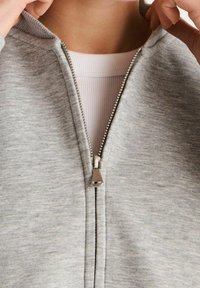 Světle šedá mikina na zip vyrobená z měkké látky, s viditelným kovovým zipem a žebrovaným vnitřním límcem. Bunda má volnější střih.