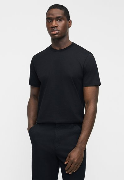 Νεαρός άνδρας με σκούρο δέρμα, φορώντας απλό μαύρο κοντομάνικο μπλουζάκι και μαύρο παντελόνι, στέκεται με το ένα χέρι στην τσέπη, μπροστά σε λευκό φόντο.