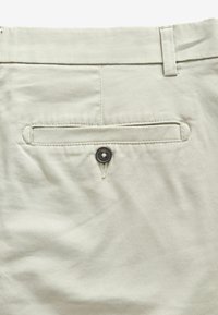 Pantalons en tissu beige clair avec une poche arrière dotée d'une fermeture à bouton noir et d'un subtil détail de couture.