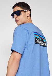 Tricou din bumbac albastru, cu un logo mare negru, având grafice cu munți și dungi colorate pe spate. Persoana poartă ochelari de soare întunecați.
