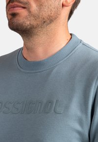 Sudadera gris con cuello acanalado y logotipo texturizado 'OSSIGNOL' en la parte delantera, confeccionada con tejido suave y acabado liso.