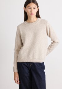 Lichtbeige sweater van zacht materiaal, met een ronde halslijn en een ribgebreide zoom met visgraatpatroon. Gecombineerd met donkere spijkerbroeken.