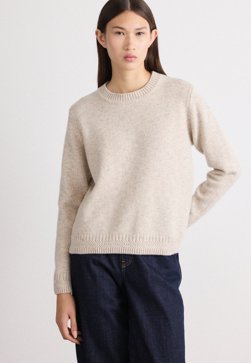 Lichtbeige sweater van zacht materiaal, met een ronde halslijn en een ribgebreide zoom met visgraatpatroon. Gecombineerd met donkere spijkerbroeken.