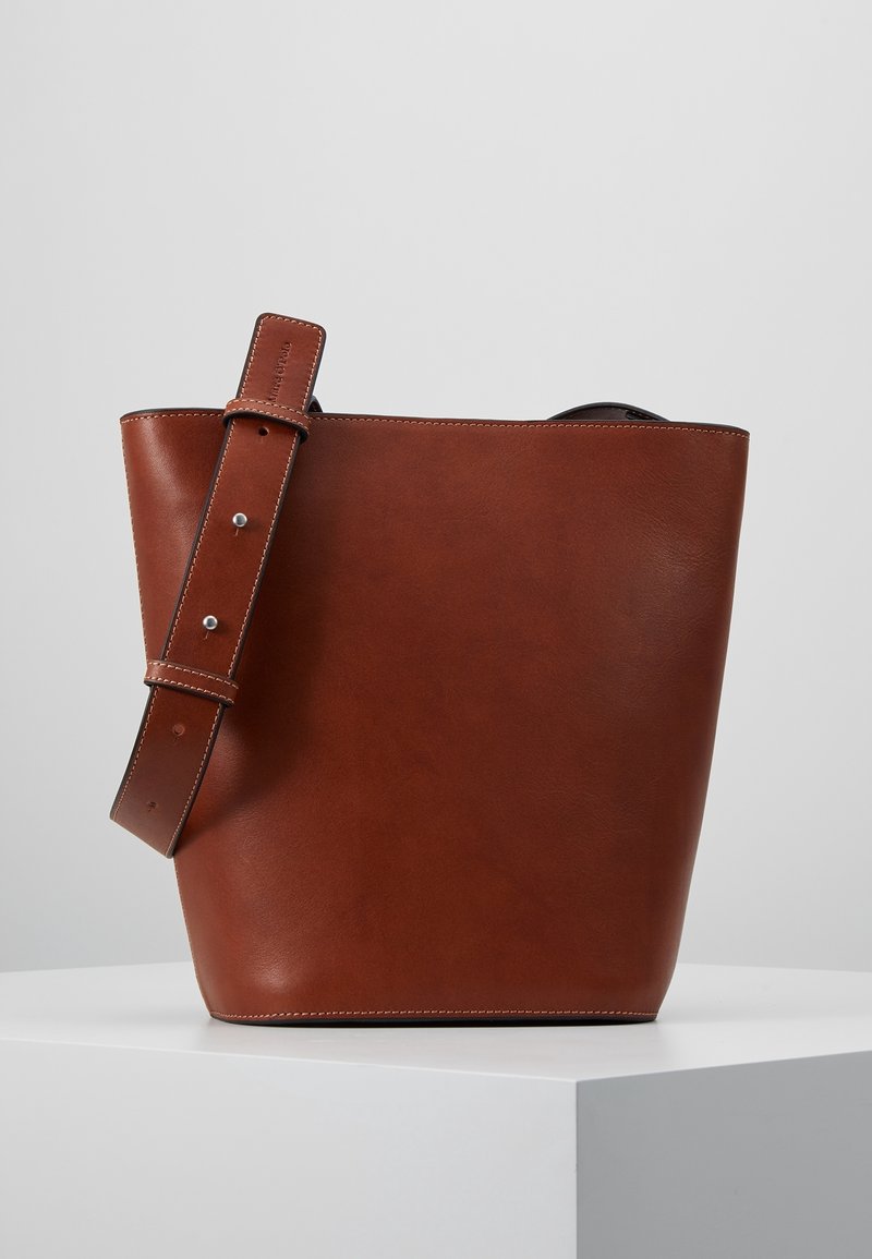 Sac seau en cuir marron avec une large bandoulière ajustable, coutures visibles et finition lisse, posé sur une plateforme blanche sur fond gris.