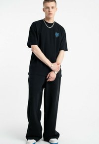 Czarna, oversize koszulka z niebieską grafiką na piersi, w połączeniu z czarnymi spodniami szerokimi i białymi sneakersami z niebieskimi akcentami, noszoną przez modela.