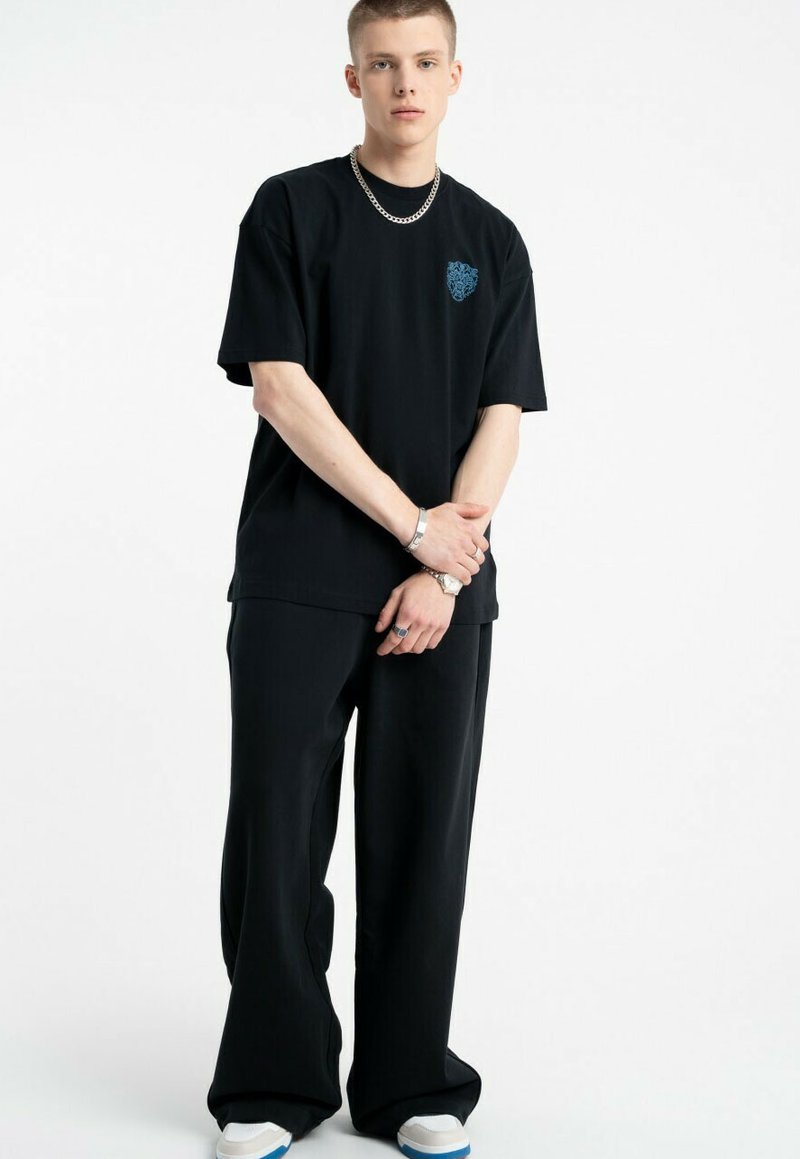 Czarna, oversize koszulka z niebieską grafiką na piersi, w połączeniu z czarnymi spodniami szerokimi i białymi sneakersami z niebieskimi akcentami, noszoną przez modela.