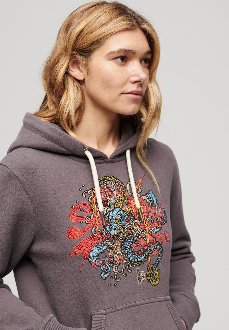 Kobieta z blond włosami ubrana w szary hoodie z kolorowym wzorem smoka i czerwonym tekstem, trzyma ręce w kieszeniach, patrzy w prawo.