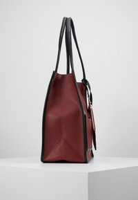 Borsa tote alta bordeaux con rifiniture nere e doppi manici, in posizione verticale su una superficie bianca contro uno sfondo semplice.