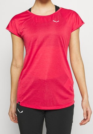 Frau trägt ein hellpinkes, kurzärmliges Sportshirt mit kleinem weißem Logo auf der Brust und schwarze Sporthose mit weißem Logo.