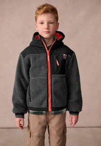 Boy wearing black and gray fleece jacket with orange zipper and hood, tan cargo pants, standing against plain background.