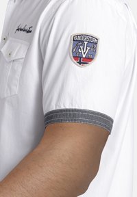 Weißes Hemd mit grauem gestreiften Bündchen und einem Patch am linken Ärmel mit "Vanderstorm 1984" und einem Anker-Design.