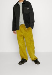 Giacca in corda nera con tasca sul petto, t-shirt grigia sotto, e pantaloni in corda gialli con tasche cargo. Scarpe nere con suole spesse.