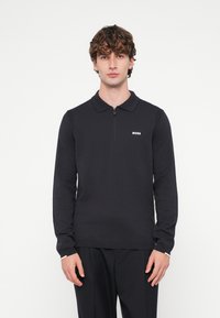 Melnas garām piedurknēm polo kreklā ar rāvējslēdzēja apkakli, ribotā tekstūra, smalks logotips krūšu rajonā, kombinēts ar tumšām biksēm.