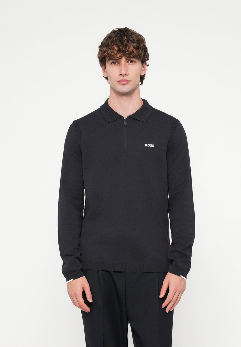 Melnas garām piedurknēm polo kreklā ar rāvējslēdzēja apkakli, ribotā tekstūra, smalks logotips krūšu rajonā, kombinēts ar tumšām biksēm.