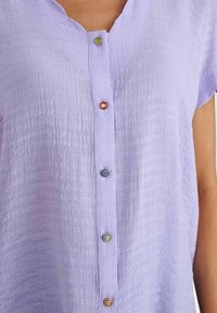 Camicia tessuta color lavanda con maniche corte e una fila verticale di cinque bottoni decorativi colorati sul davanti.