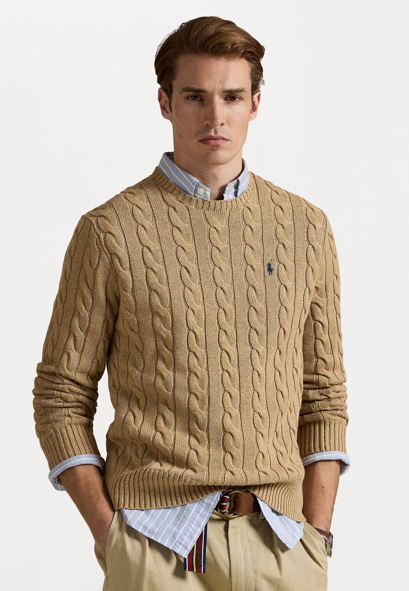 Beige kabelgebreide trui met een ronde hals, gecombineerd met een lichtblauw overhemd met kraag. De trui heeft ribgebreide manchetten en onderkant.