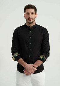 Chemise noire à col mandarin, avec des poignets à motifs floraux. Le tissu semble lisse avec une coupe structurée.