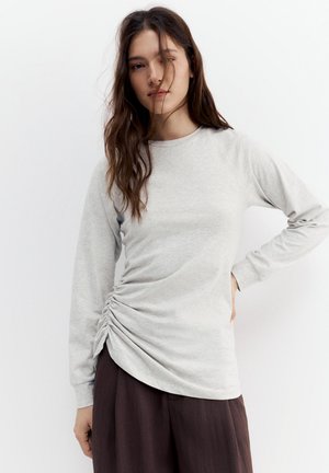 Žena s dlhými hnedými vlasmi, oblečená v svetlošedej nariasenej blúzke s dlhými rukávmi a voľných tmavohnedých nohaviciach, stojaca s bielym pozadím.