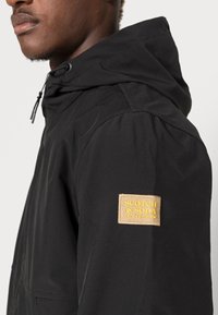 Giacca nera con cappuccio e finitura testurizzata, caratterizzata da una patch con logo ricamato in oro sulla manica sinistra e cordini regolabili.