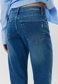 Siniset farkut, joissa on keskikorkea vyötärö. Takana näkyvä etiketti ja oikeassa taskussa salaman muotoinen yksityiskohta.