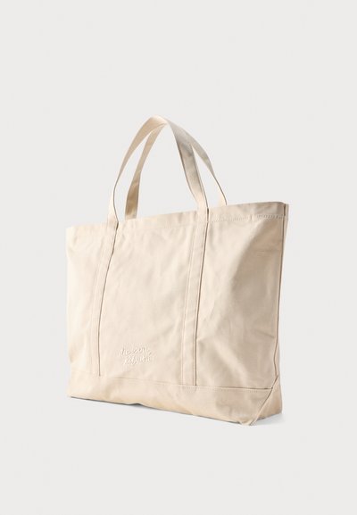 Μπεζ τσάντα καμβά με διπλές λαβές, ορθογώνιο σχήμα και λεπτό κεντημένο λογότυπο στο κέντρο μπροστά.