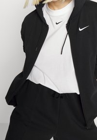 Personne portant un T-shirt blanc Nike sous un sweat à capuche noir Nike non zippé et un pantalon noir avec une taille à cordon.